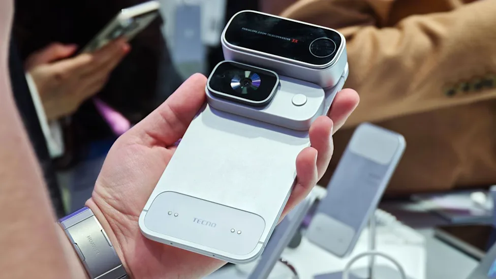A hand holding the Tecno modular phone with the action camera module and a 3x periscope zoom teleconverter stacked on the back
