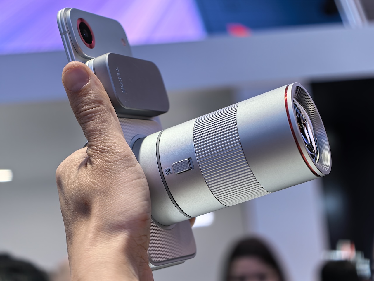 A hand holding the Tecno modular phone with the large cylindrical telephoto lens module attached, the lens barrel dwarfing the phone